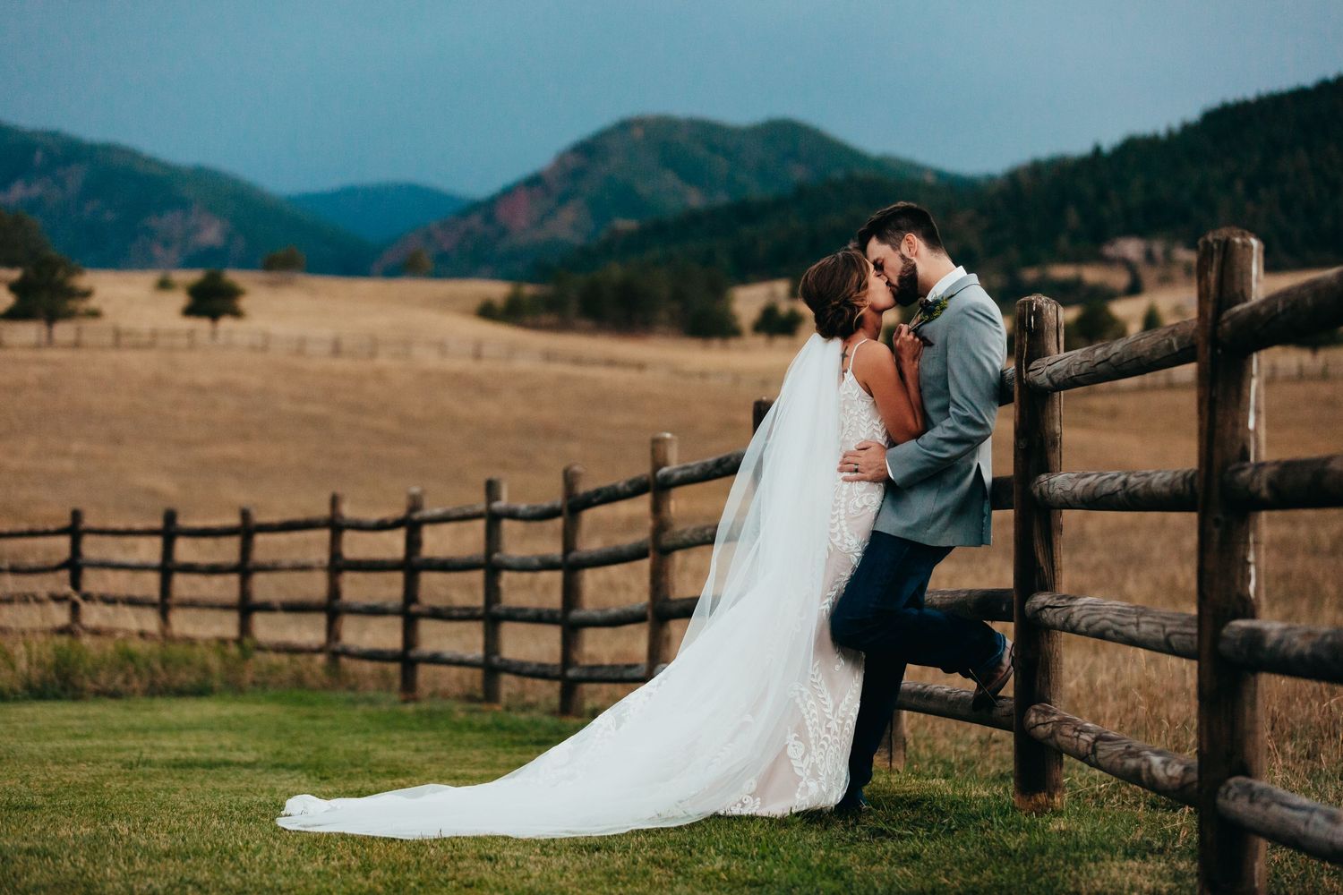 Romantic wedding portrait of couple embracing by rustic fence with mountain landscape backdrop.