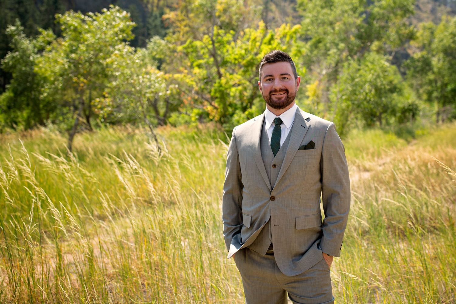 Suited groomsman poses confidently in mountain meadow with sunlit greenery in background.