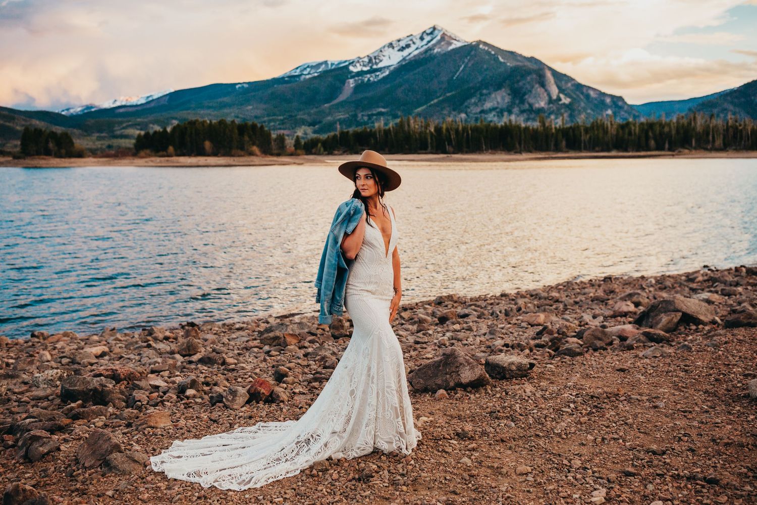 Bride in lace dress and denim jacket poses by mountain lake with snow-capped peaks.