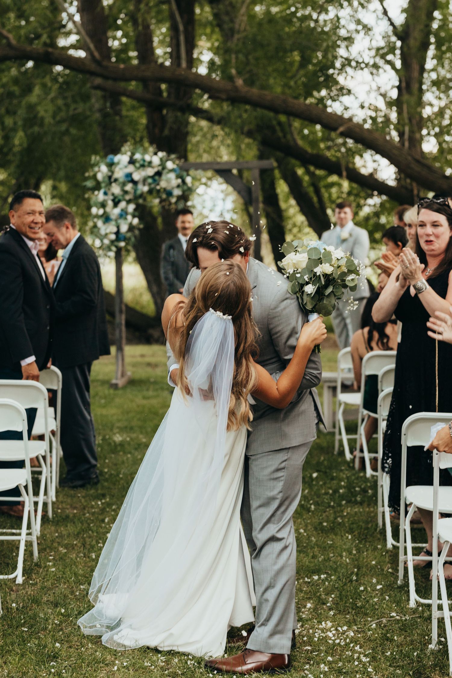 Romantic wedding ceremony kiss in an outdoor garden setting with white chairs and floral arch.