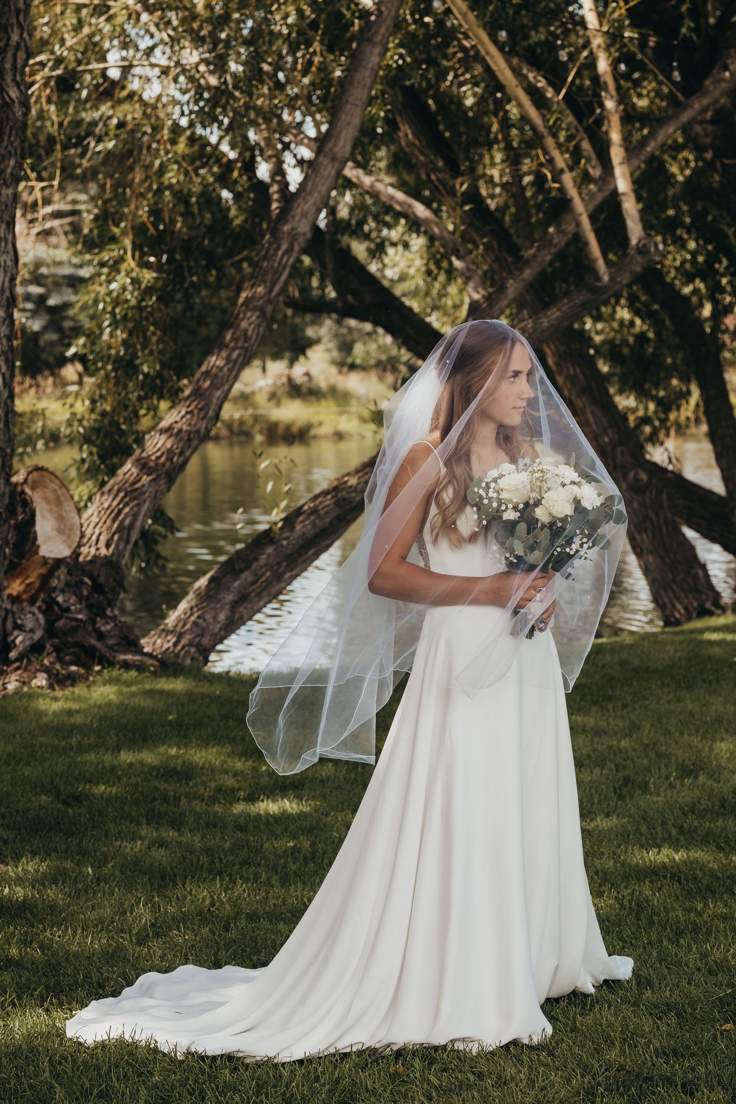Bride in flowing white dress holds bouquet near water during outdoor wedding portraits.