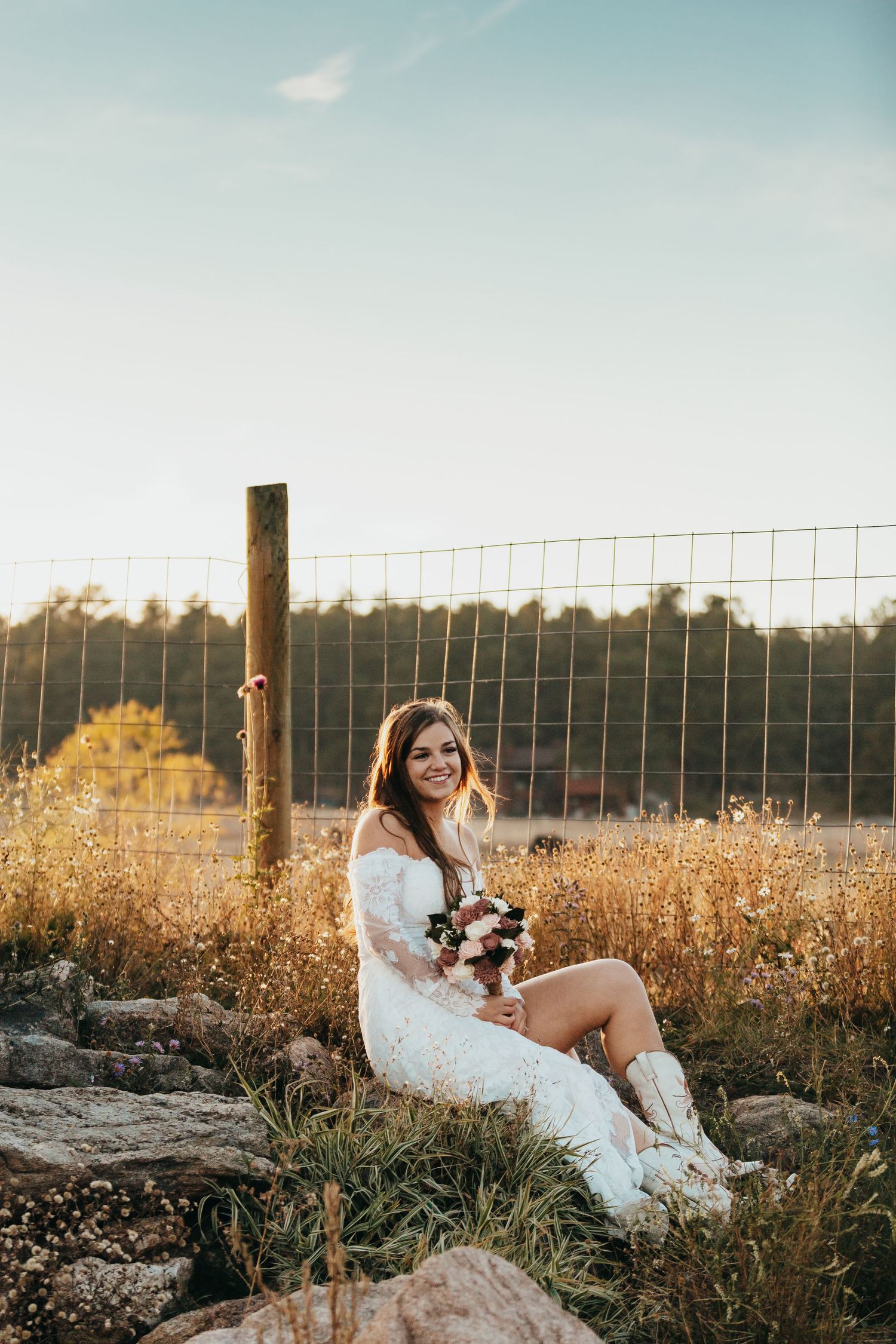 Person in white dress sitting on rocks at sunset with rural fence in background.