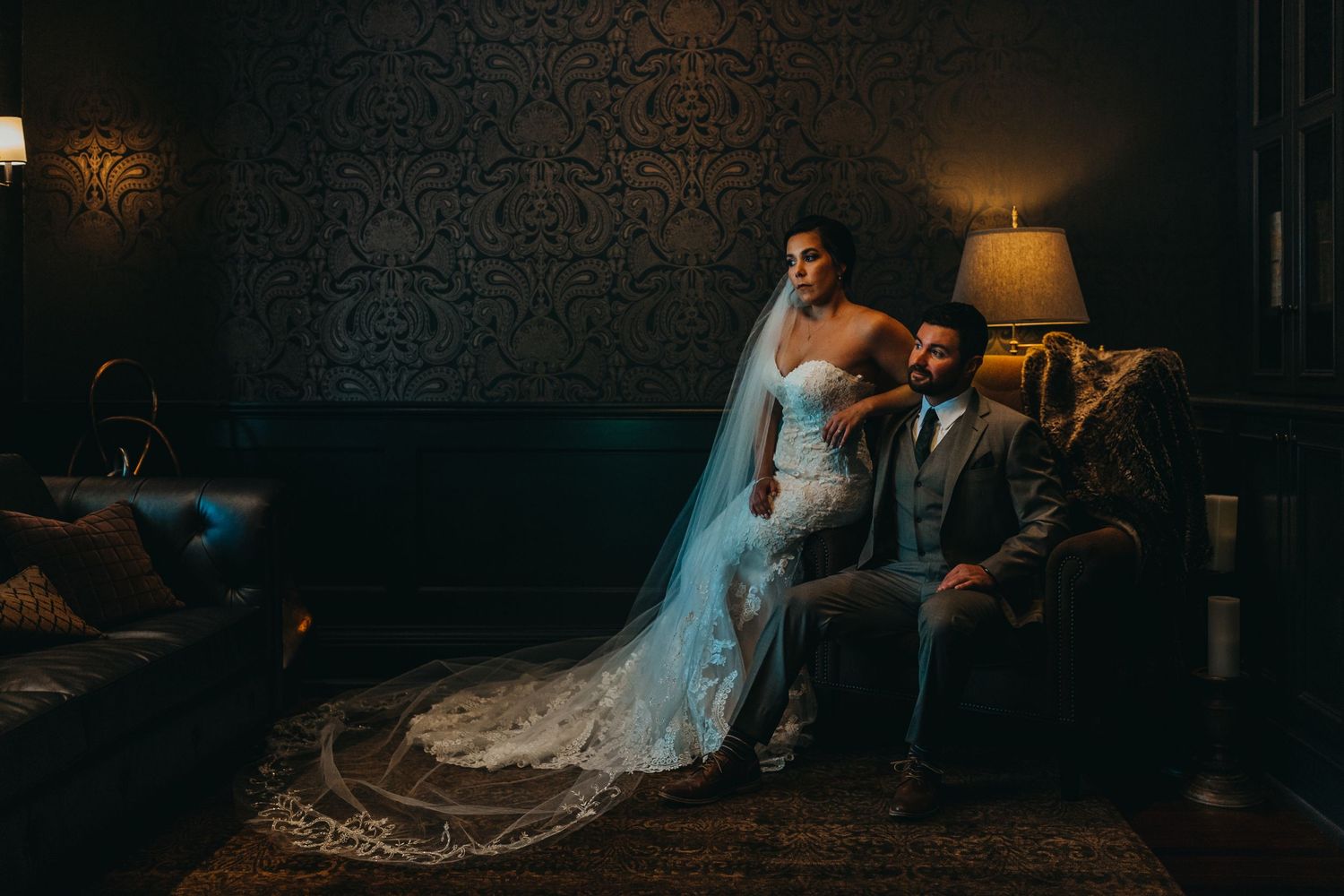 A wedding couple seated on a vintage couch in a dimly lit room with ornate wallpaper.