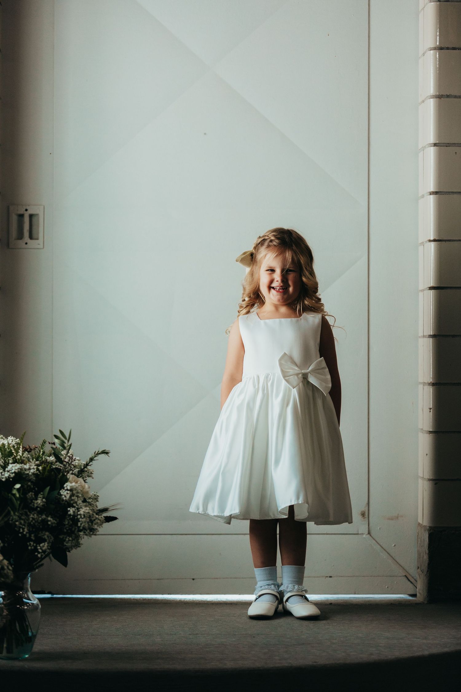 A happy child in white dress with bow standing near wall.