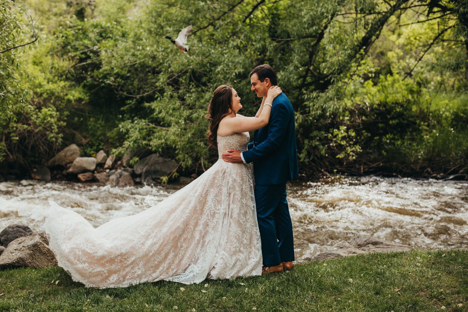 Couple in formal attire embrace by rushing river with lush greenery.