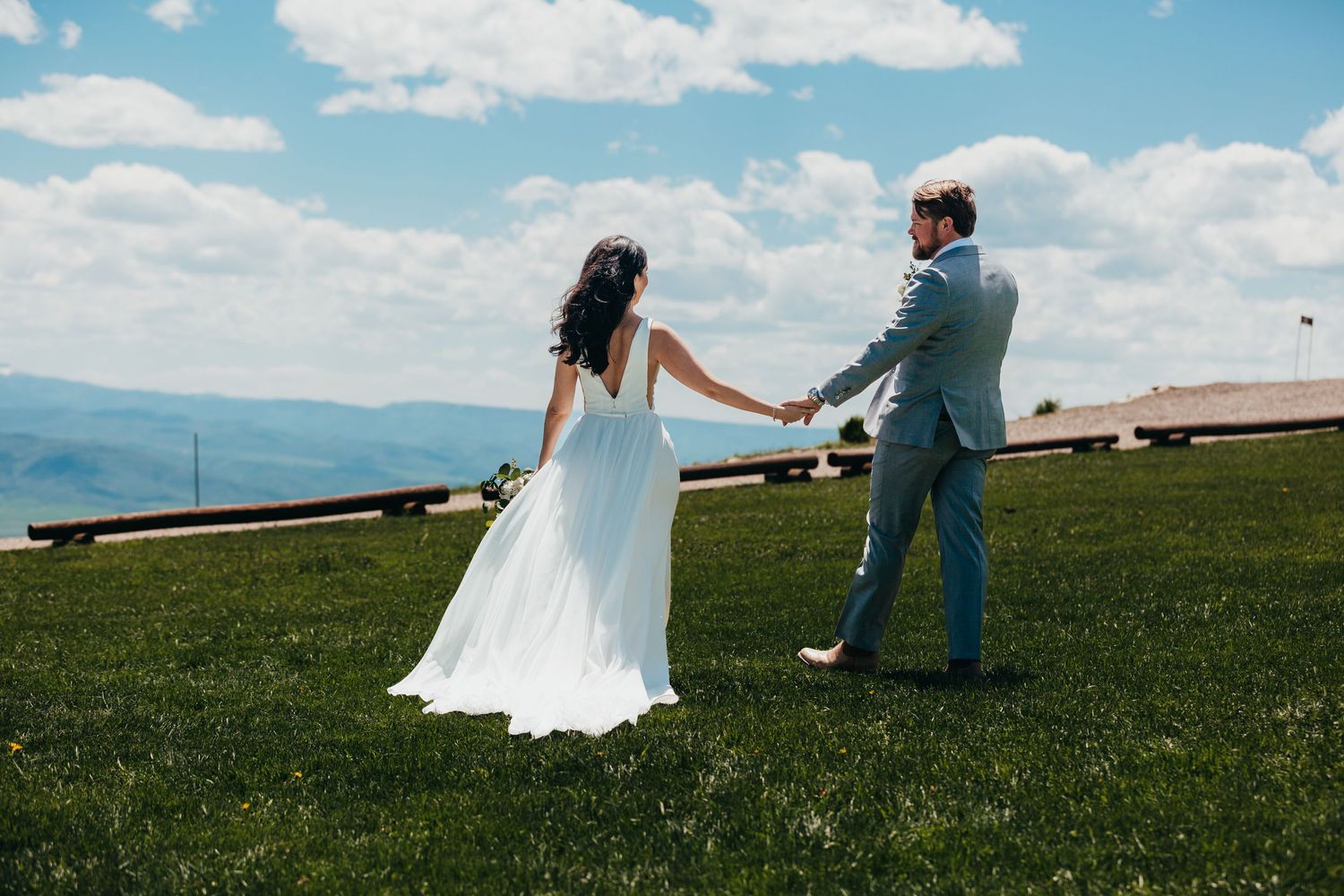 Couple dancing on grassy hilltop with mountains and blue sky in background.