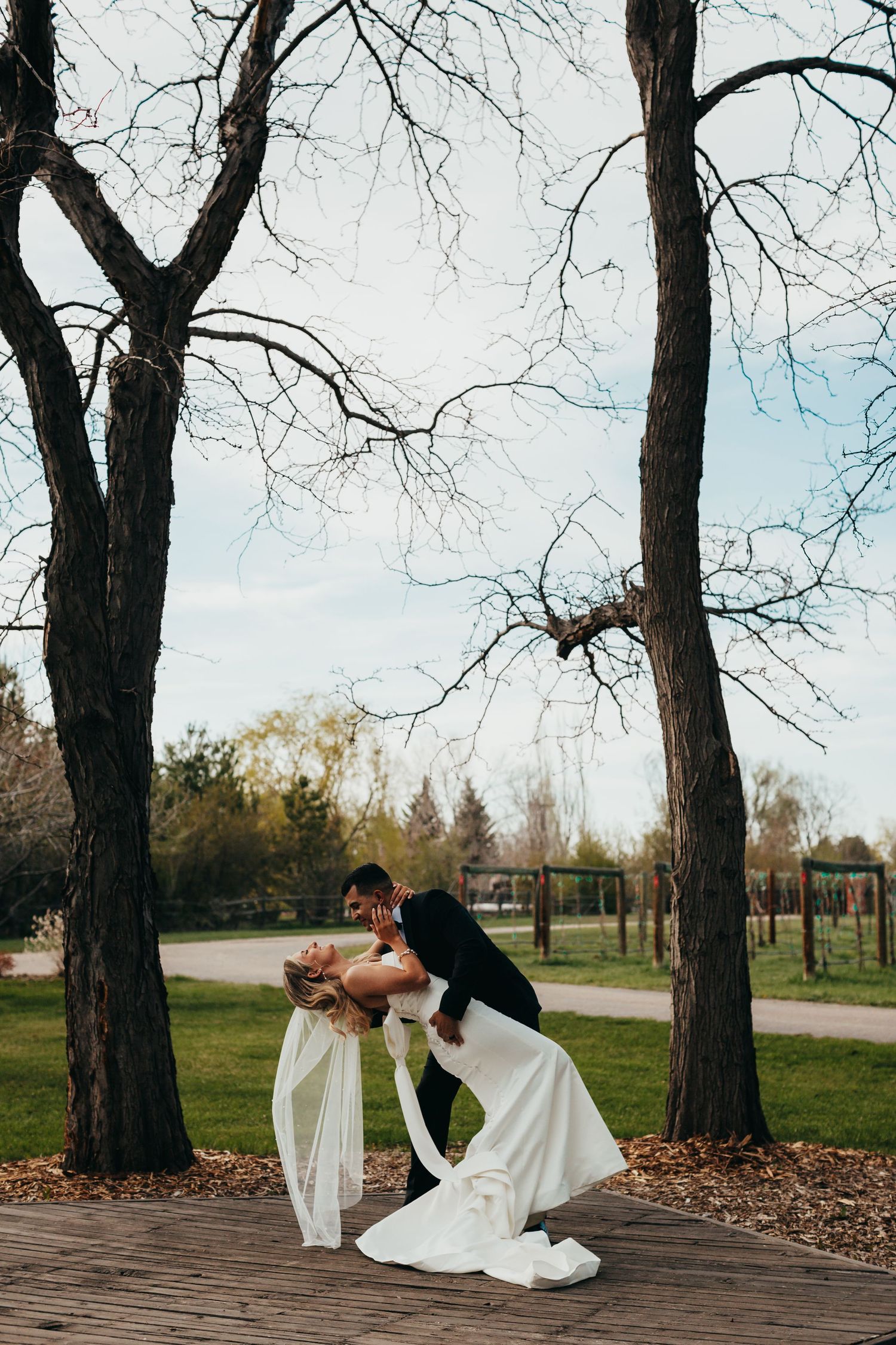 Two dancers in formal attire perform a romantic dip on an outdoor wooden deck surrounded by bare trees.