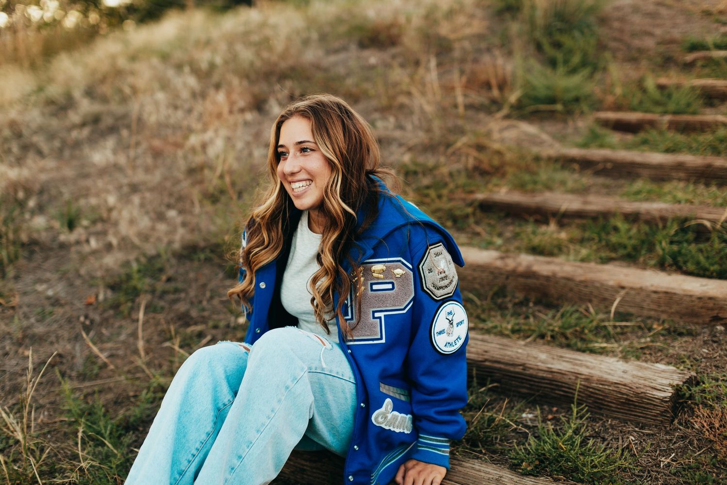 Person wearing varsity jacket with patches sitting outdoors on rustic steps.
