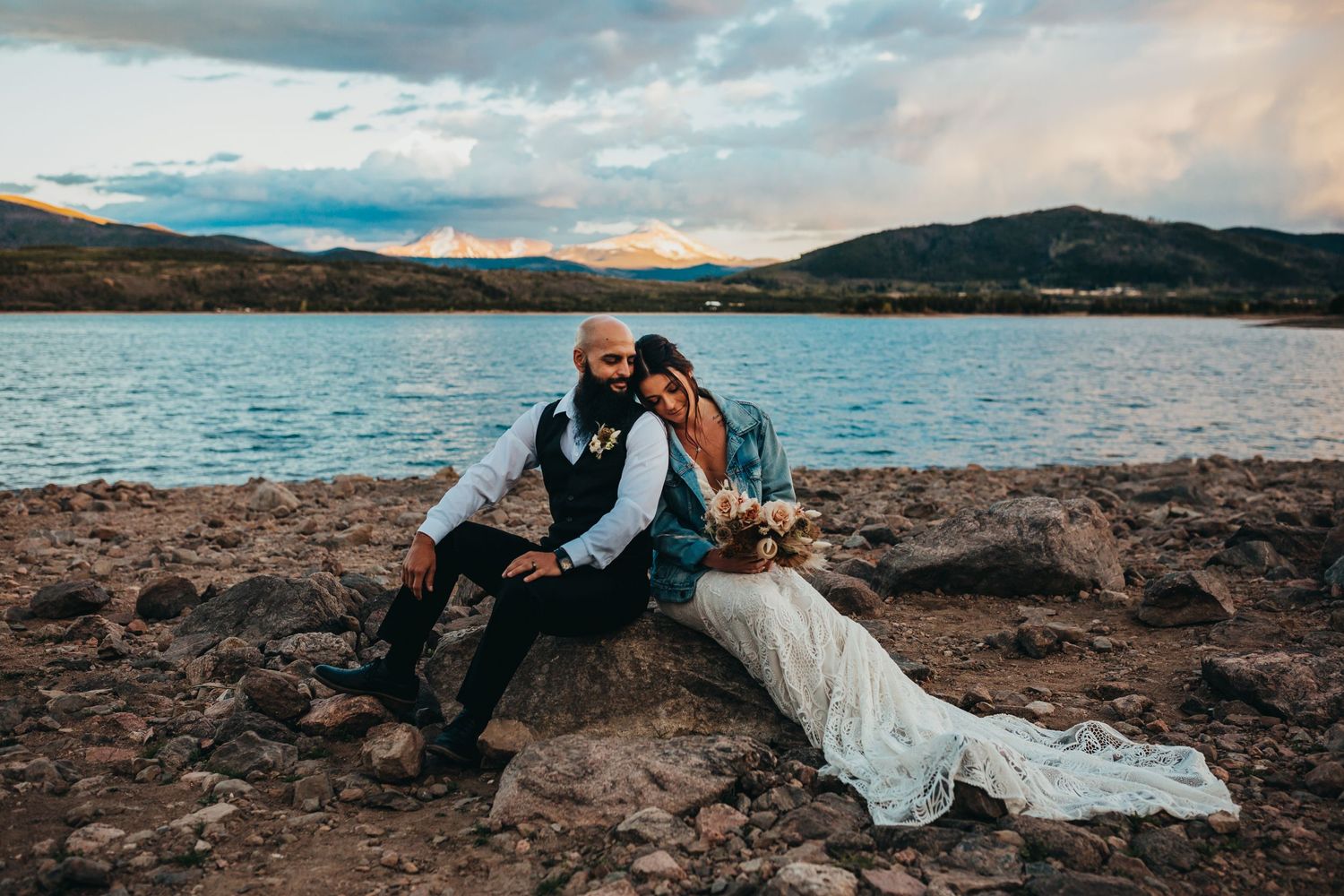 Wedding couple in formal attire sitting on rocks by lakeside during sunset with mountains in background.
