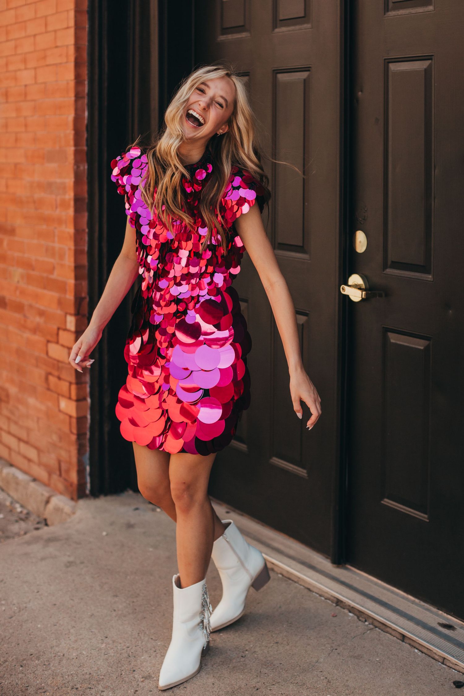 Woman in bright pink sequined dress and white boots dancing joyfully by brick wall.