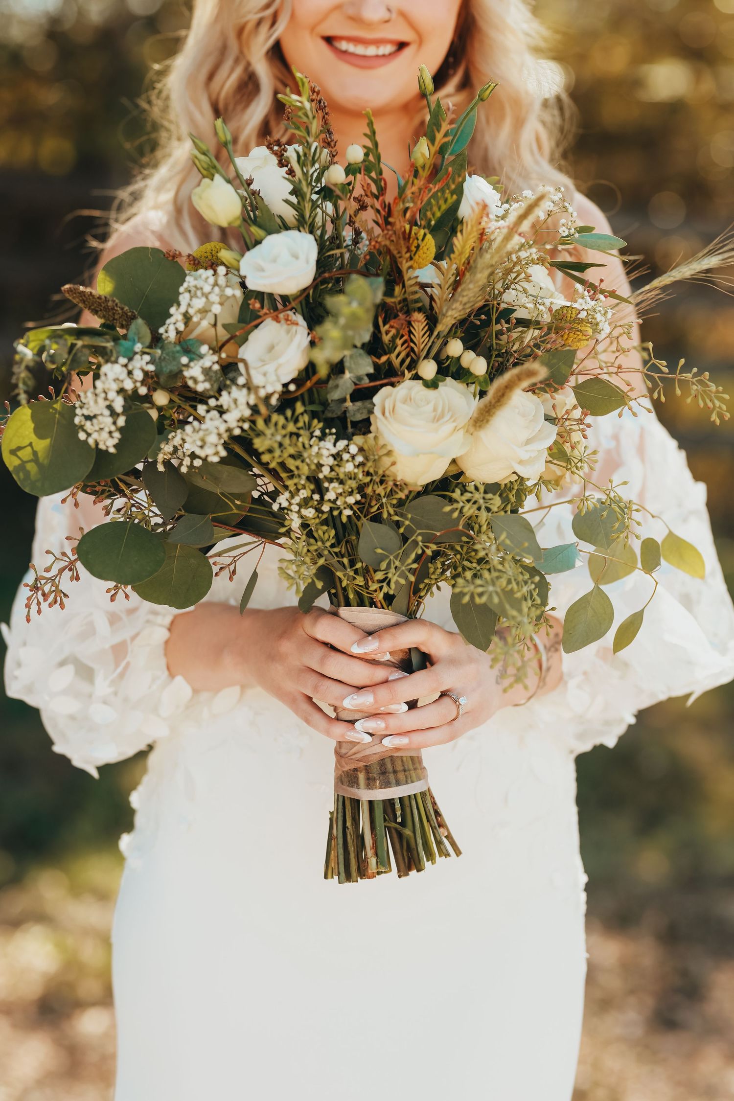 Wedding bouquet features white roses, eucalyptus, and dried elements against lace dress details.