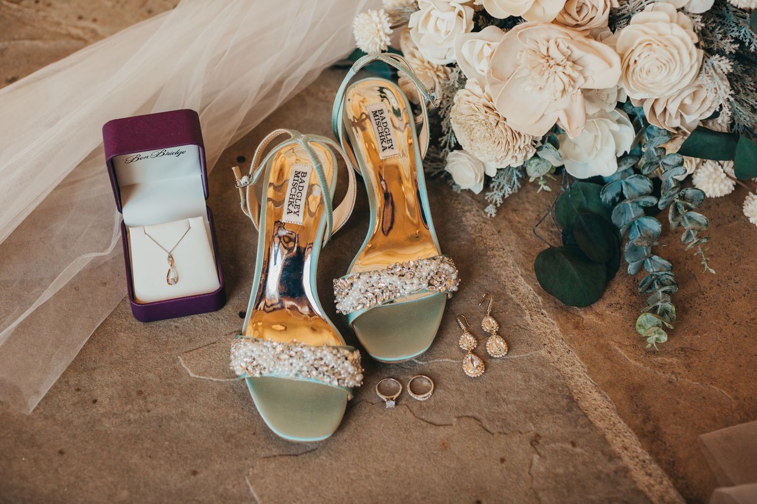 Wedding accessories including sparkly heels, rings, and white roses arranged on stone surface.
