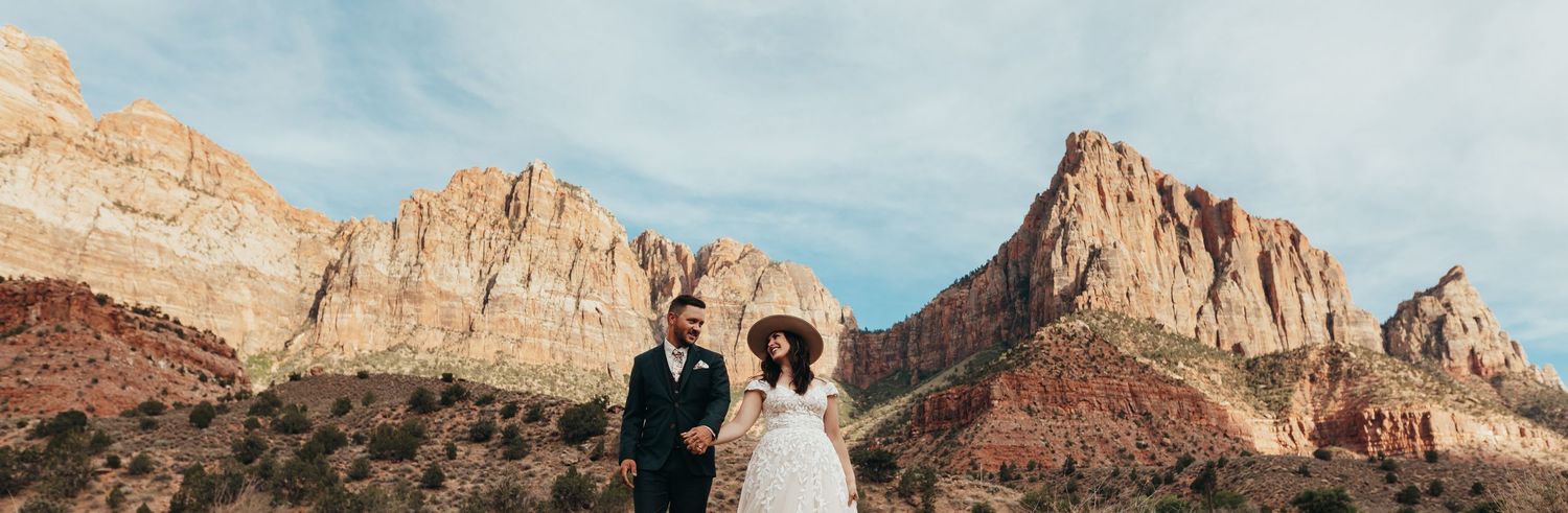Wedding couple stands beneath dramatic red rock formations in a desert landscape.