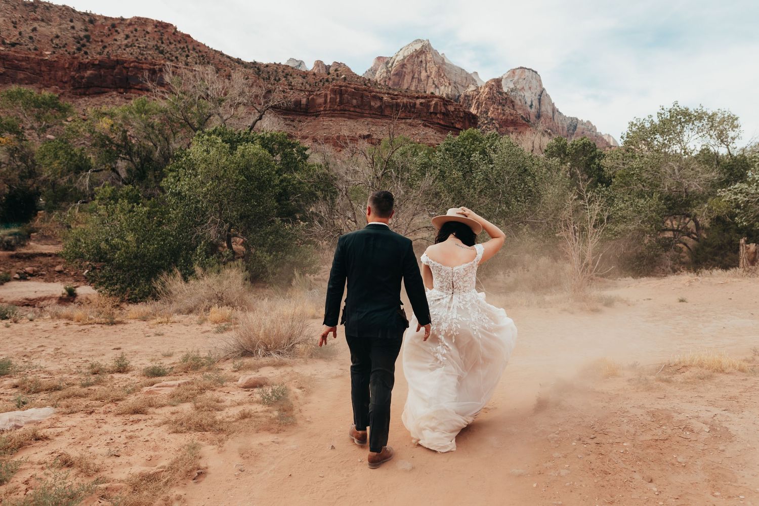 Couple walking through desert dust with majestic canyon cliffs in the background.