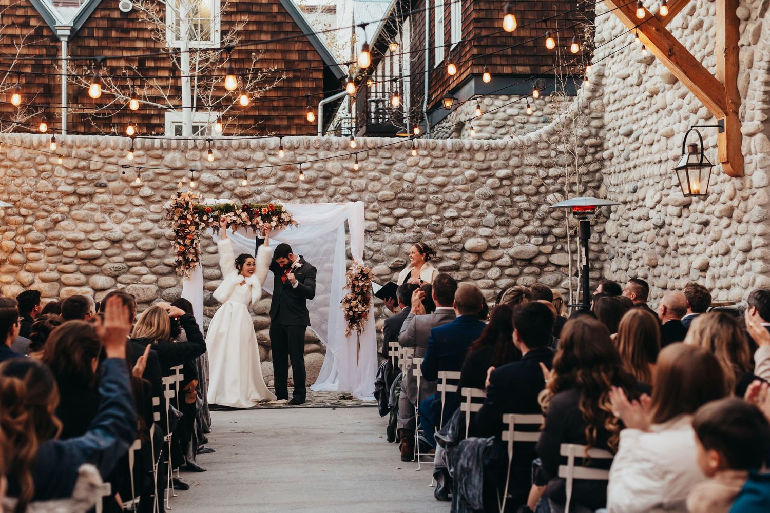 A rustic indoor wedding ceremony in a brick-walled venue with string lights and rows of seated guests.