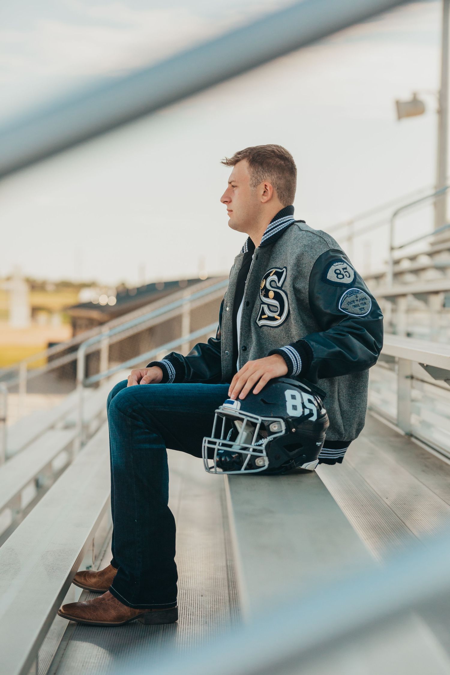 Athletic senior portrait of student in letterman jacket sitting on bleachers at sports field.