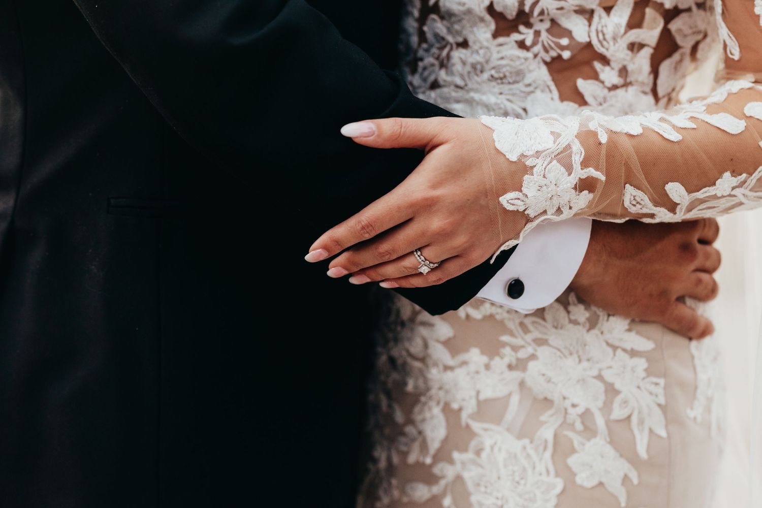 Close-up detail shot of hands embracing during a wedding ceremony showing lace sleeves and formal attire.