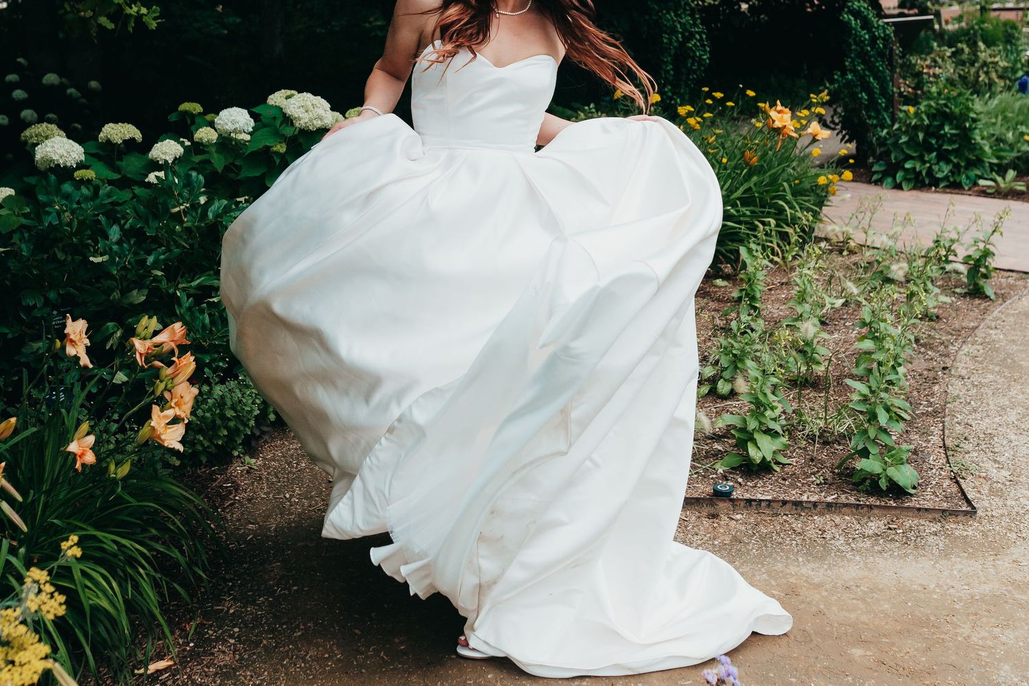 The hem of a flowing white wedding dress swirls in a garden setting.