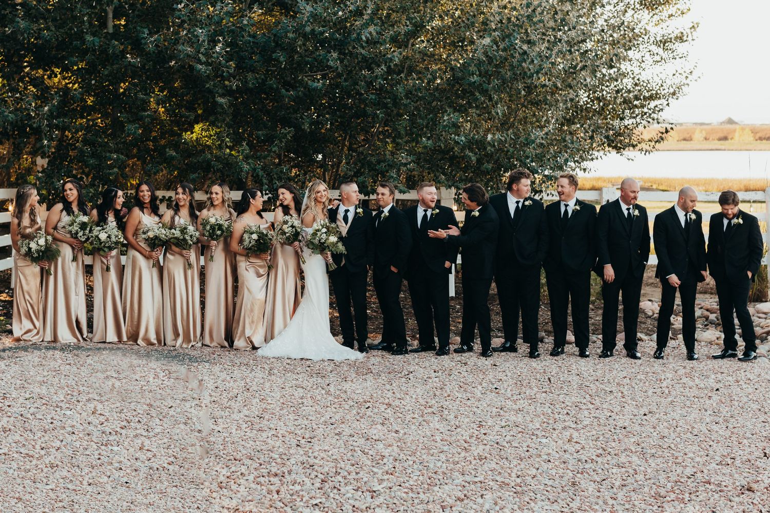 Wedding party in black suits and beige dresses poses together outdoors on gravel pathway.
