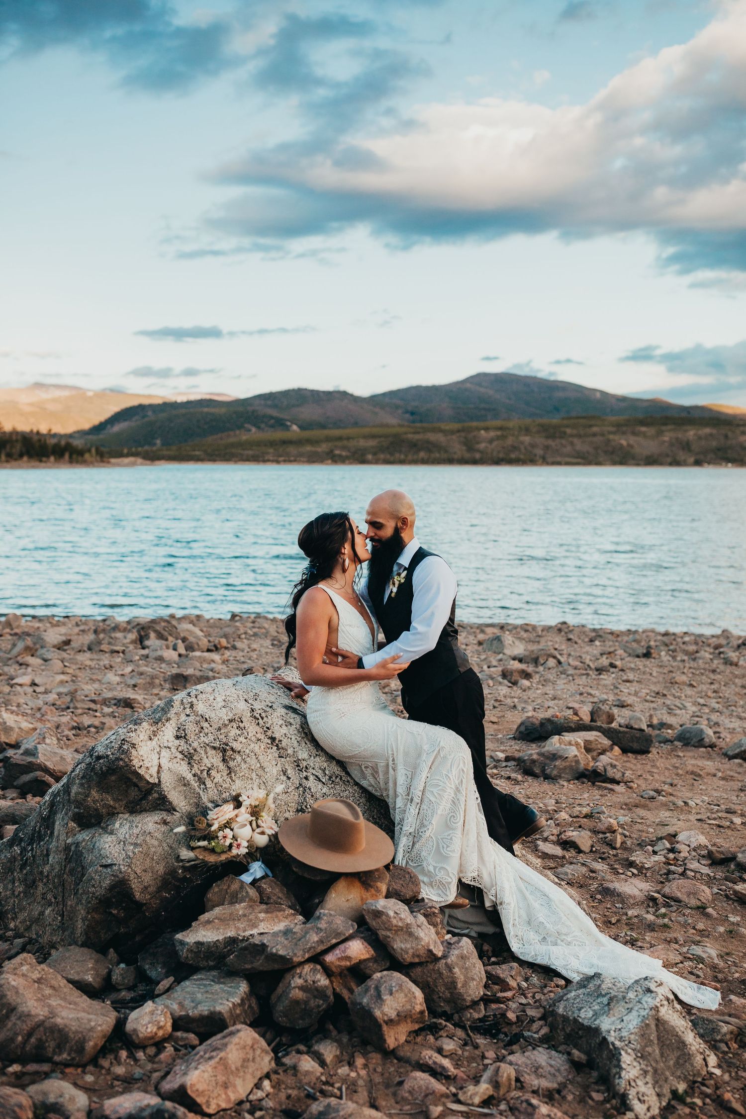 Romantic wedding photo at sunset by a mountain lake with couple sitting on rocky shore.