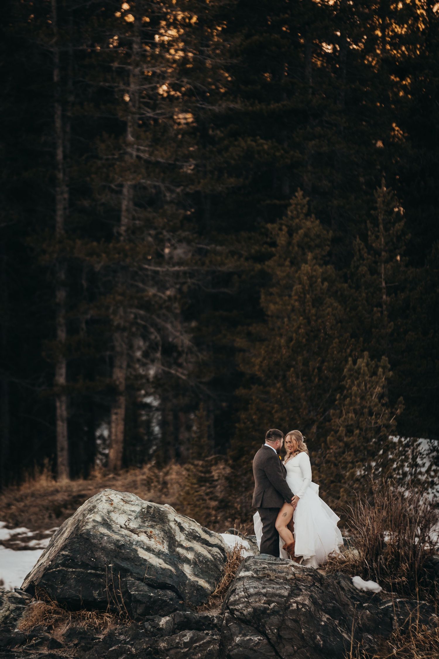 Wedding couple stands intimately on snow-covered rocks surrounded by forest.