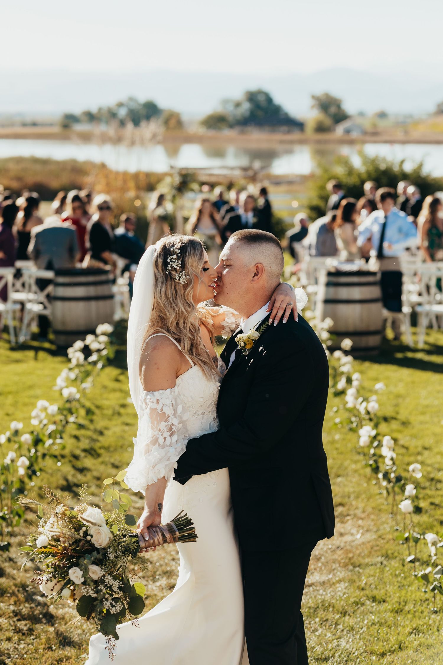 Newlyweds share their first kiss during an outdoor garden wedding ceremony with floral decorations and guests in background.