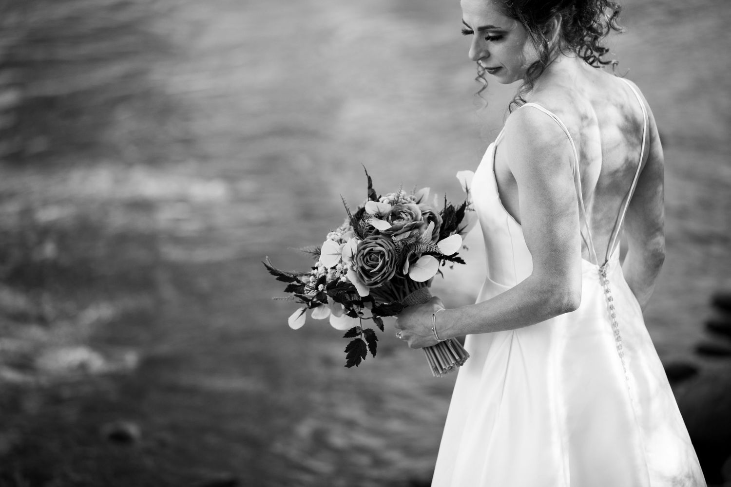 Black and white photo of a bride holding a bouquet beside a river.