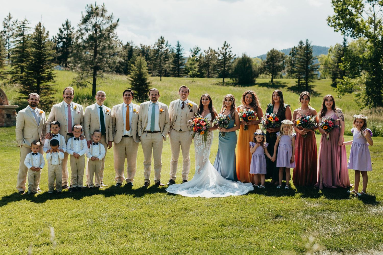 Wedding party in pastels and neutrals poses on grassy hillside with evergreen trees in background.