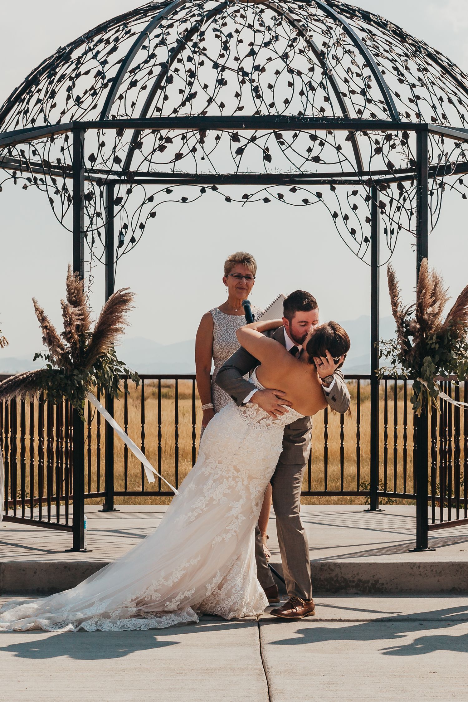 Wedding couple shares romantic dip pose under decorative metal gazebo with pampas grass decor.