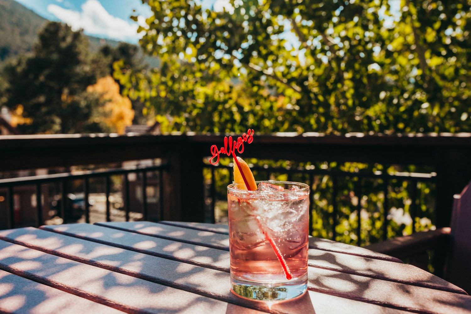 Pink cocktail with floral garnish on outdoor patio table with mountain view in background.