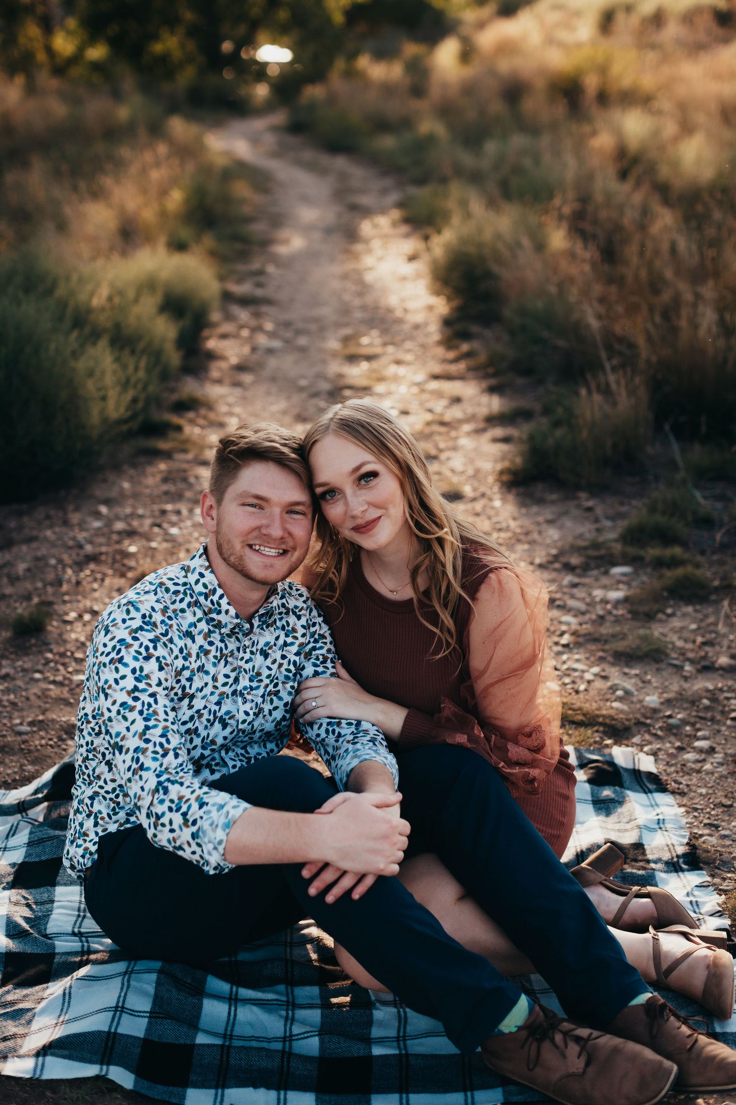 Couple sitting on plaid blanket along scenic dirt trail during sunset.