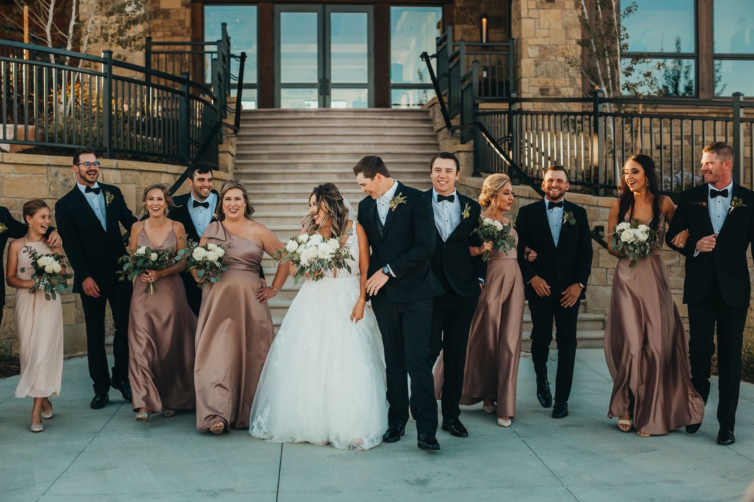 Full wedding party in formal attire poses together outside a stone building.