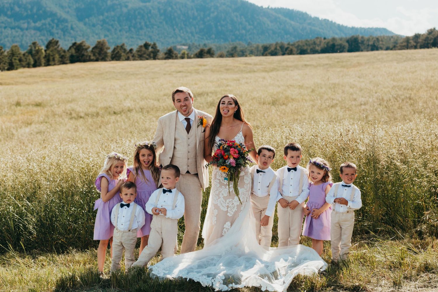 Wedding party poses in golden field with mountain backdrop and children in pastel attire.