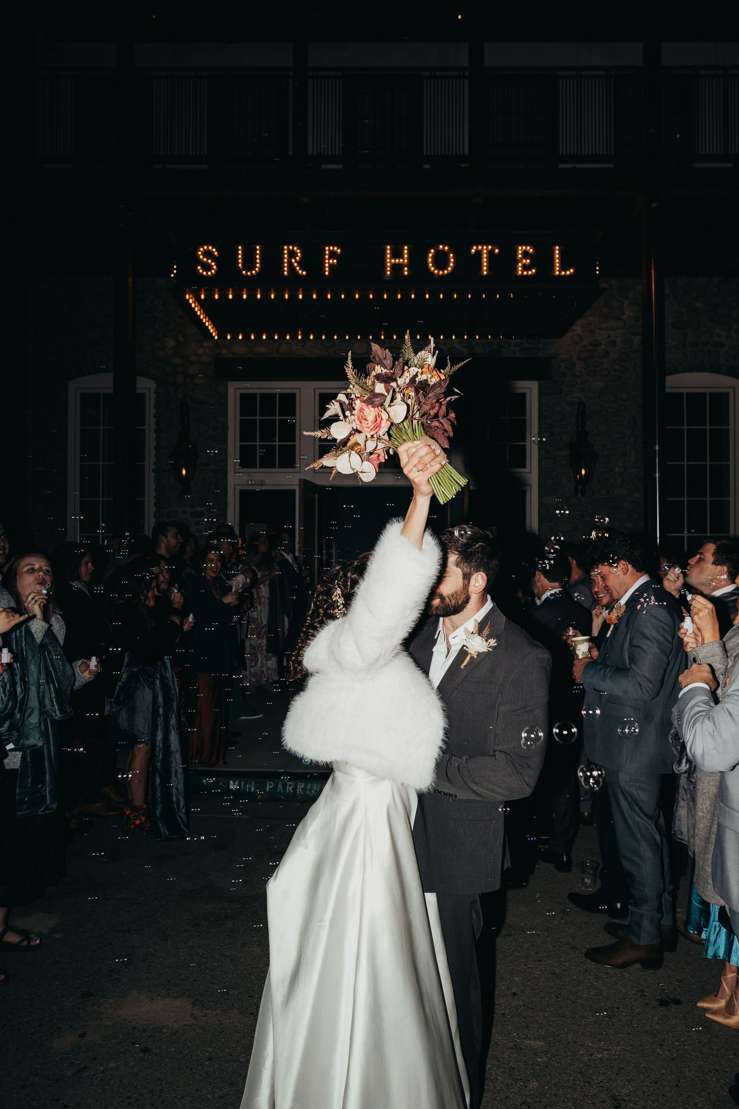 Wedding couple celebrates outside the Surf Hotel at night with the bride tossing her bouquet under illuminated signage.