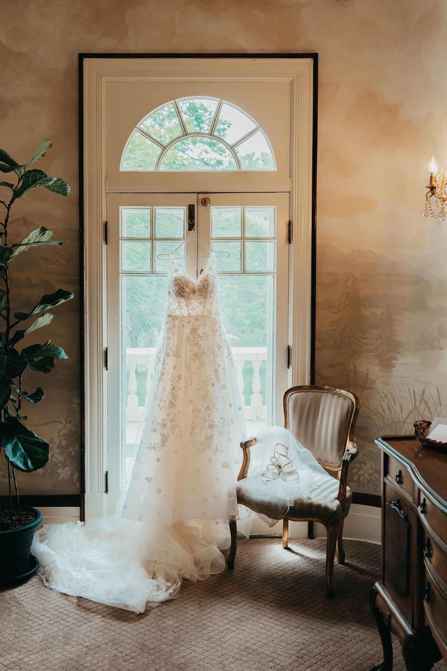 Elegant lace wedding dress hanging in window frame with antique chair.