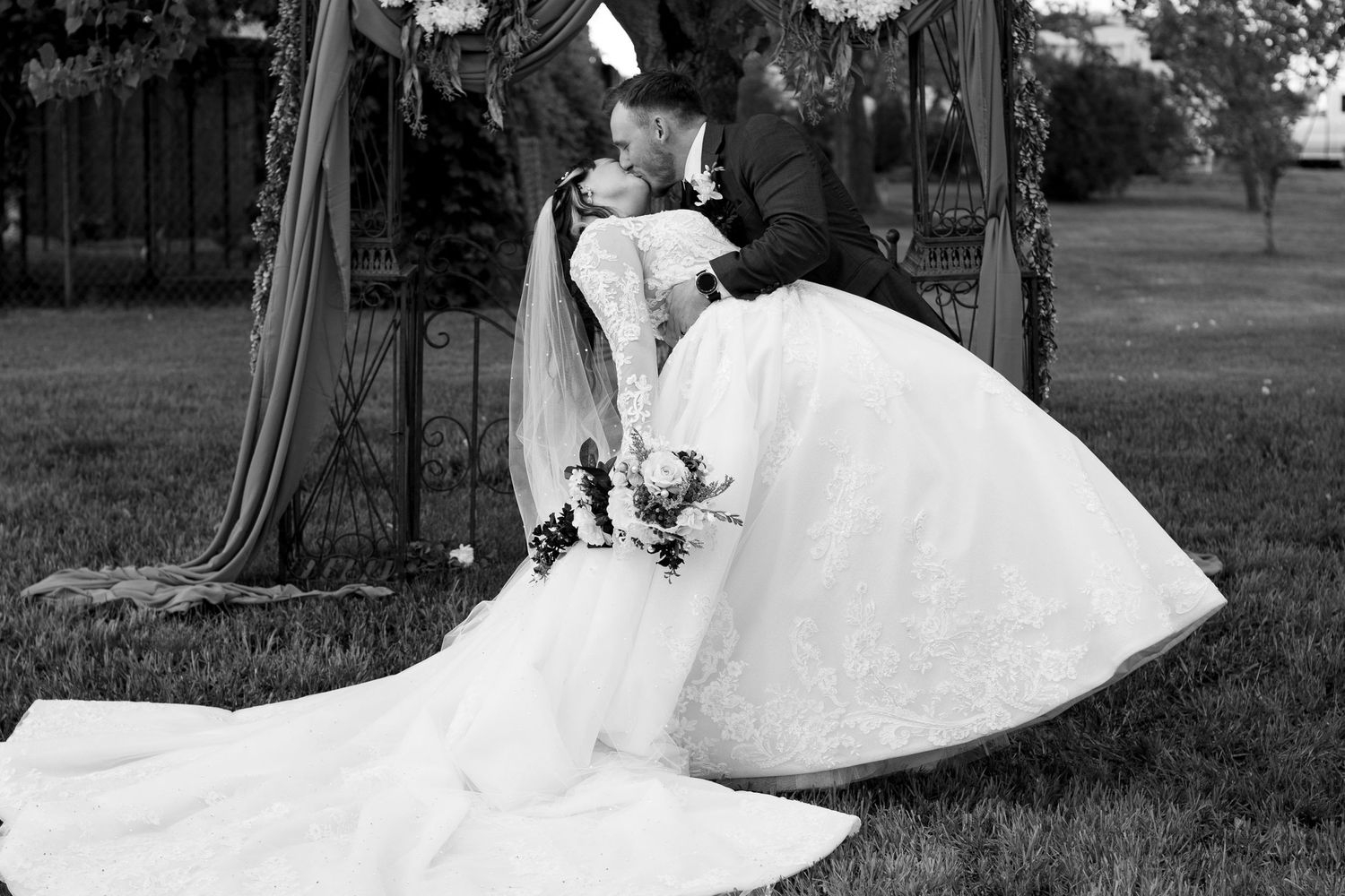 Black and white photo of a couple sharing a romantic dip pose under a wedding arch.