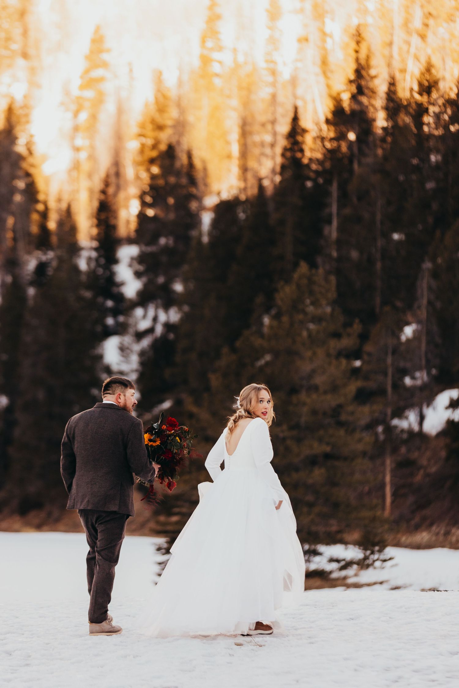 Couple walking through snowy landscape at sunset with pine trees in background.