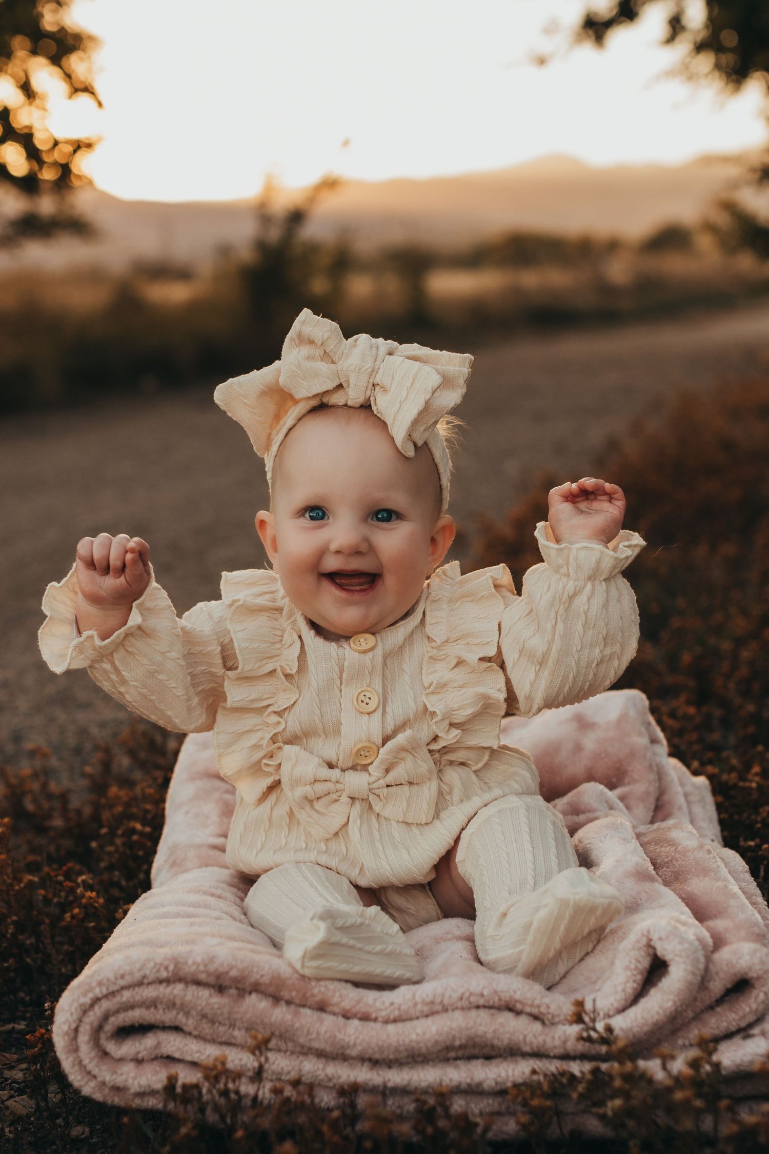 Baby in cream knit outfit with bow sits on blanket during sunset photoshoot.