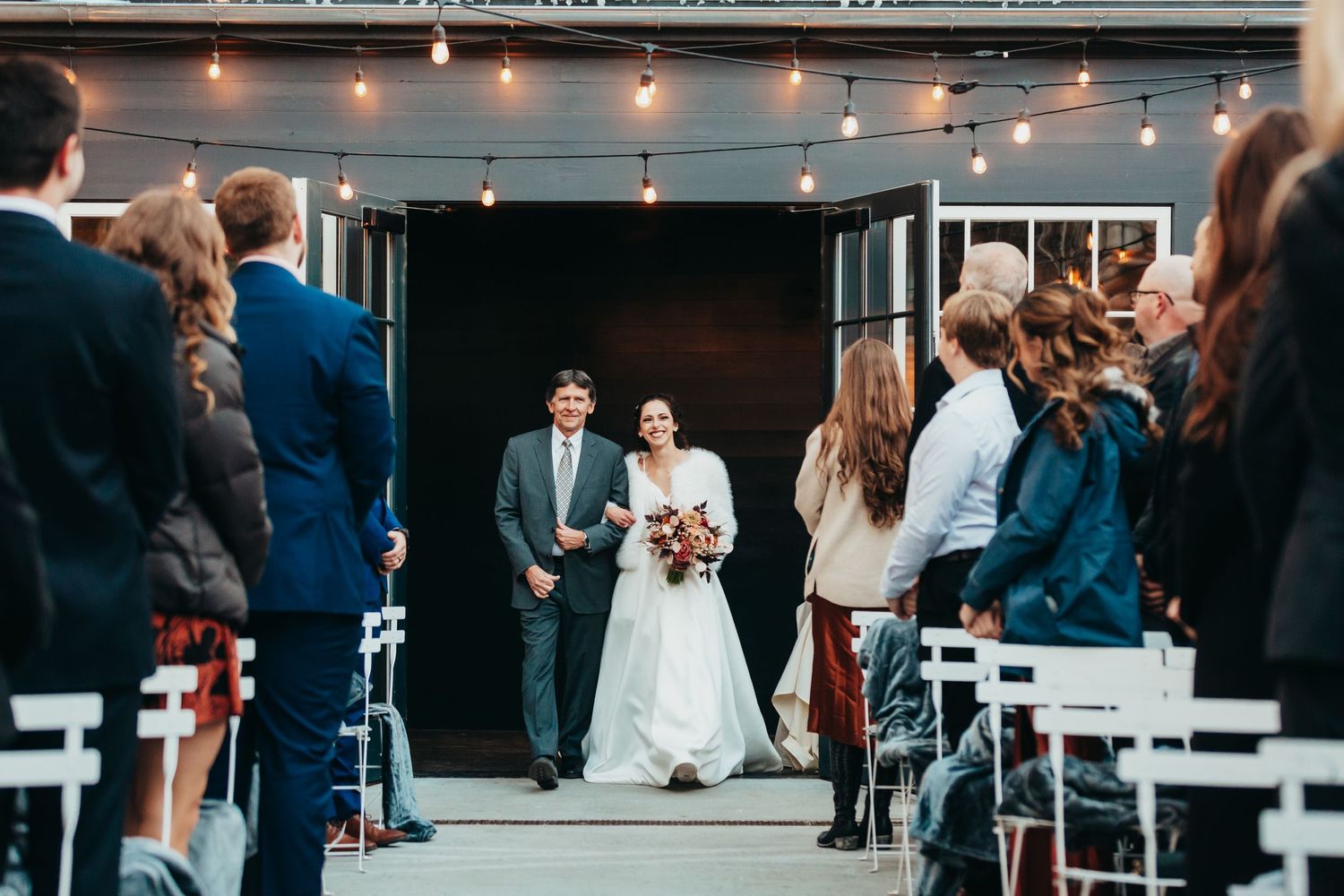 Wedding ceremony processional under string lights with guests seated in white chairs.
