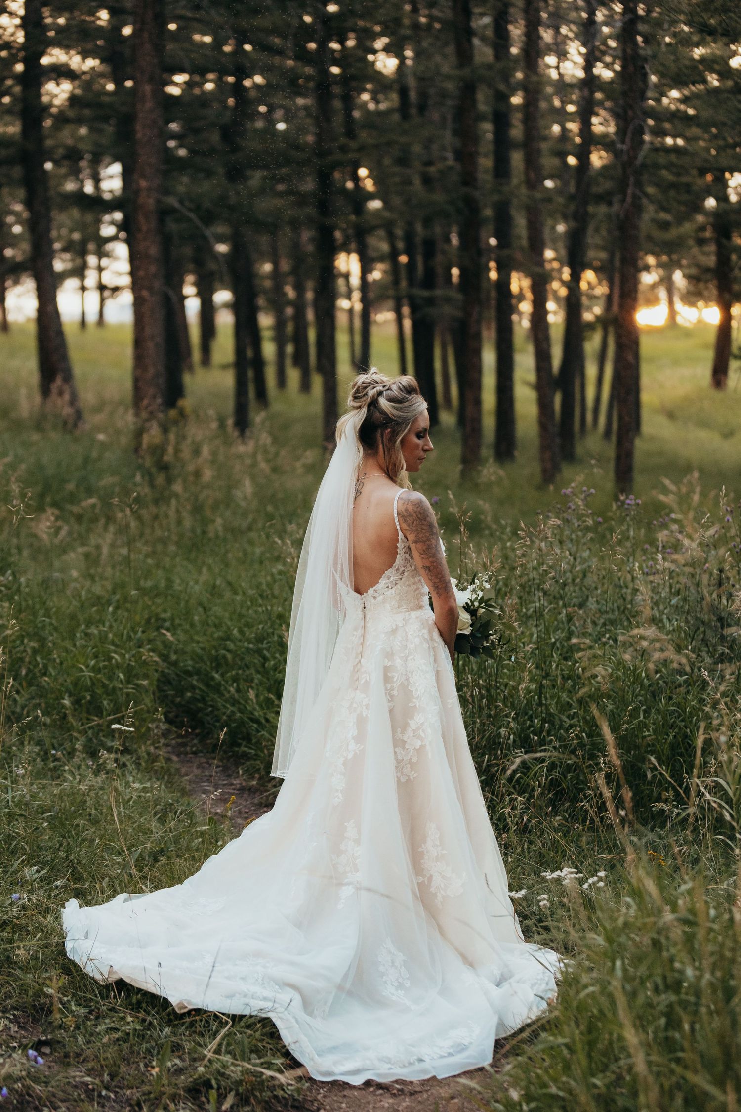 Back view of bride in lace wedding dress with long veil standing in forest setting.