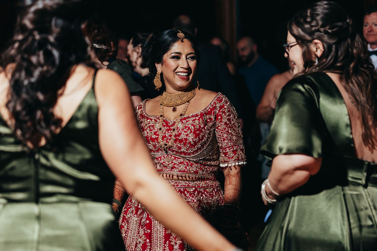 Wedding party members in green dresses celebrate with the bride wearing a red lehenga.