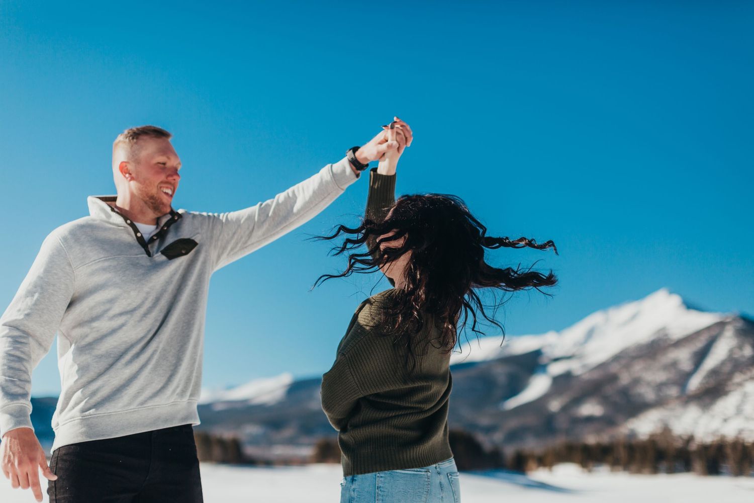 Couple dancing playfully in snow with mountain backdrop on sunny day.