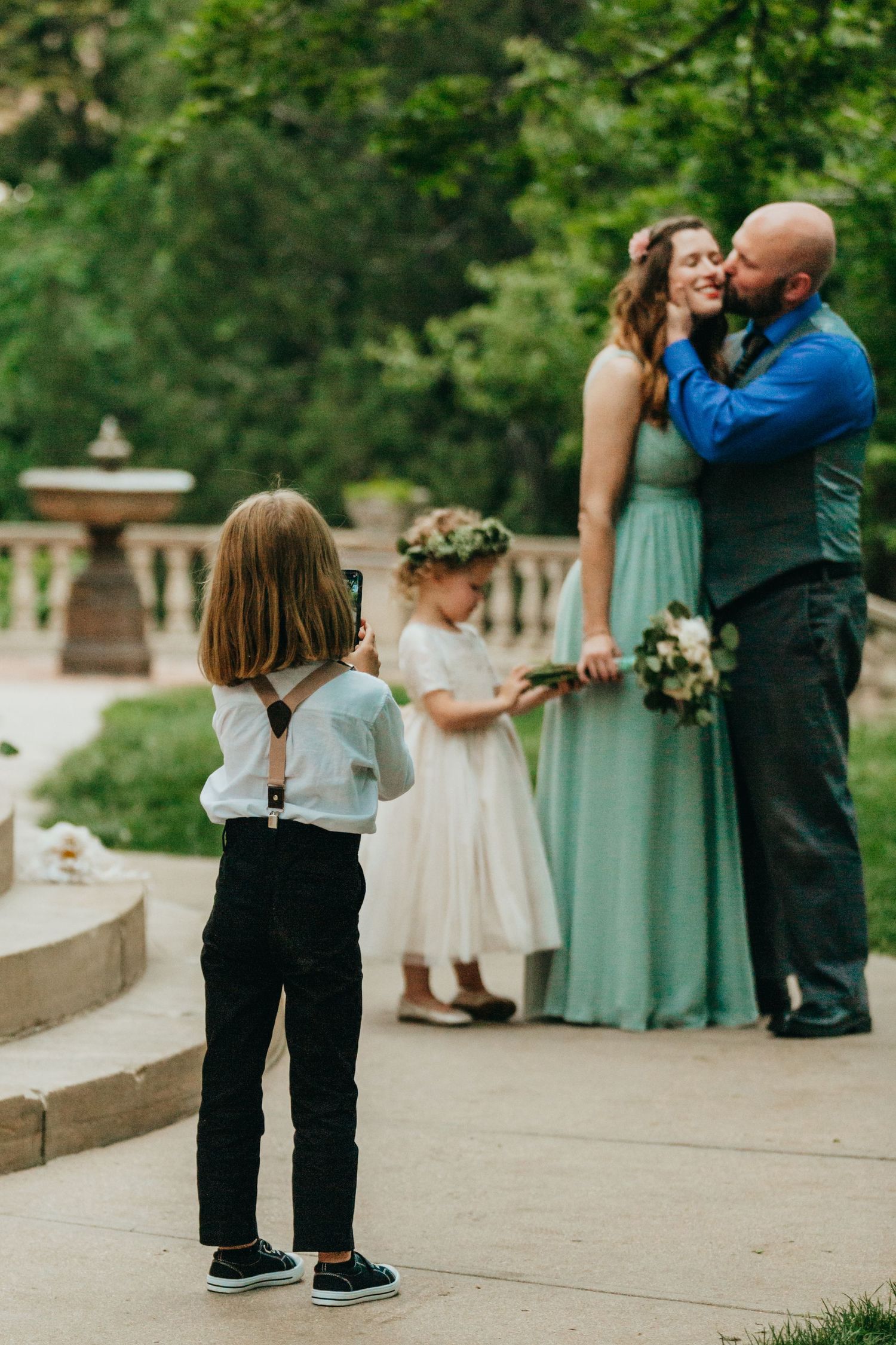 A young child photographs a wedding moment while standing on outdoor stone steps.