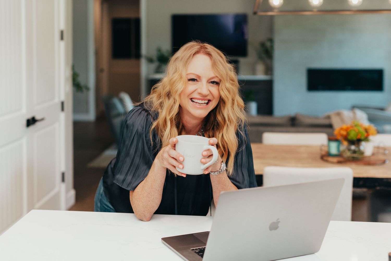 Person enjoying coffee while working on laptop in modern white kitchen.