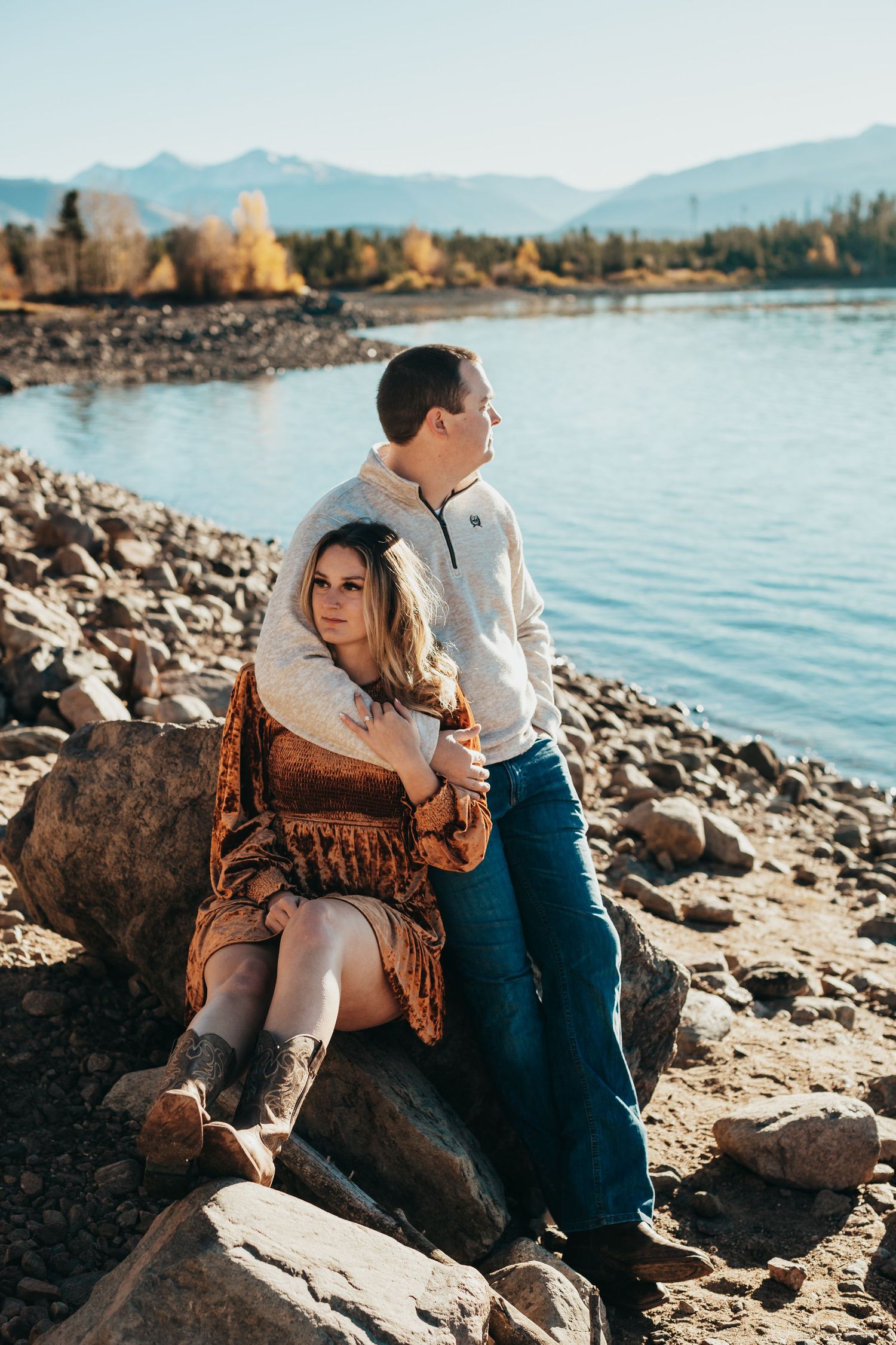 Couple embraces on rocky shoreline with mountain lake and autumn trees in background.