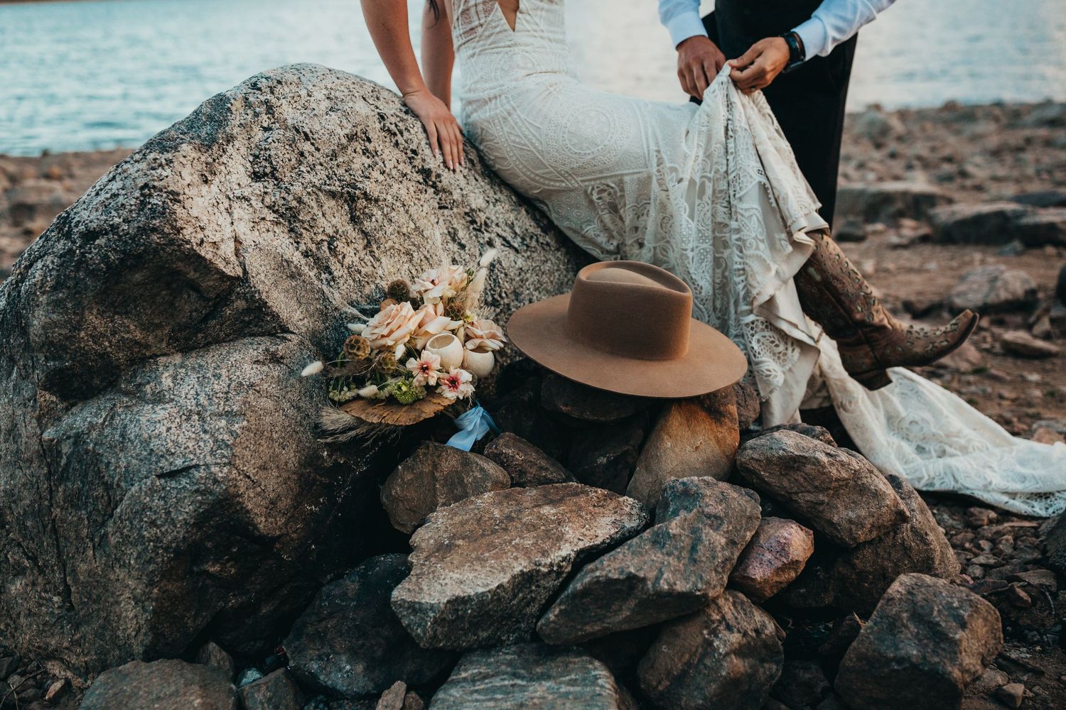 Wedding dress train and brown hat arranged on rocky shoreline with bouquet.