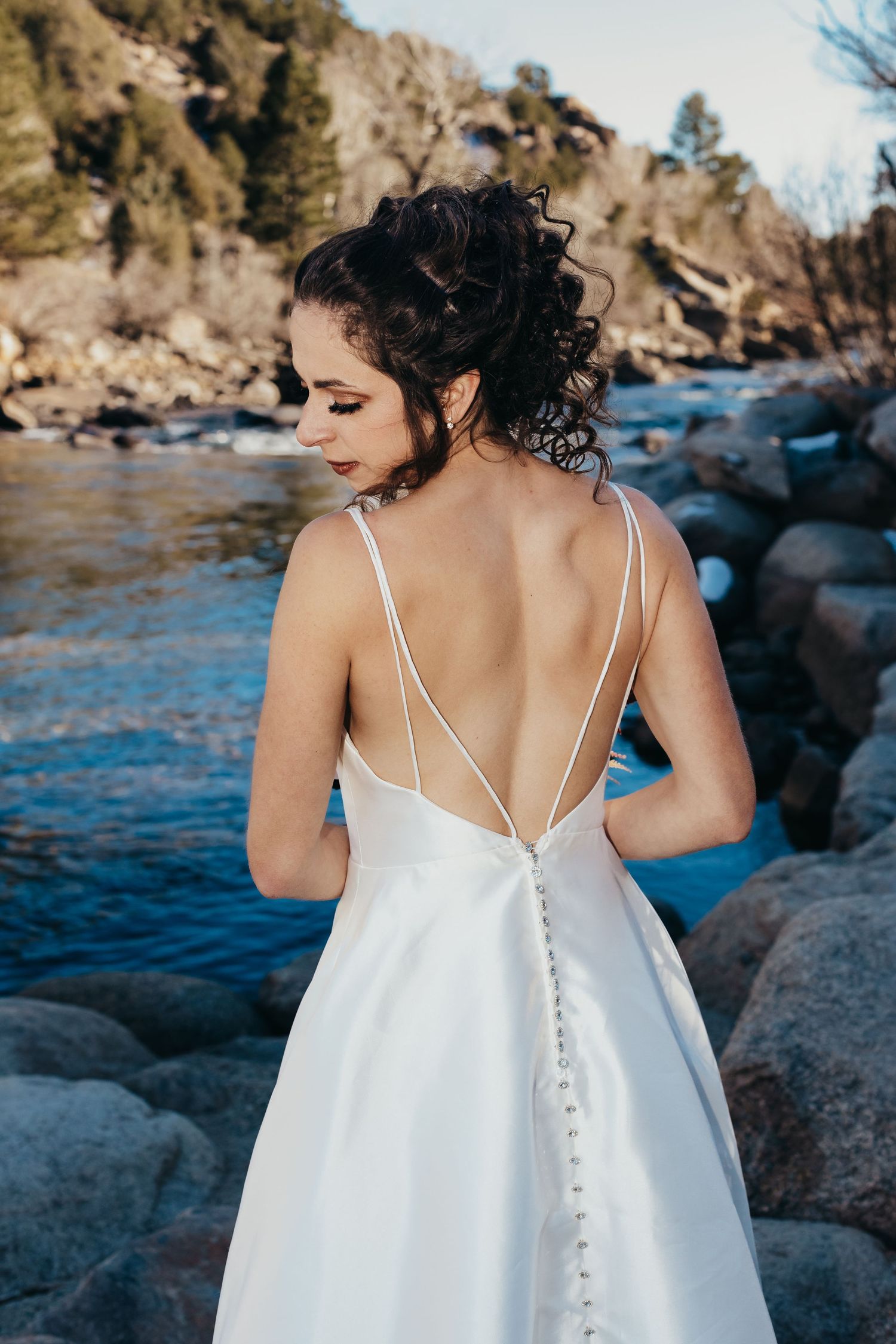 Back view of a bride in a strappy white wedding dress standing by a river with rocks.