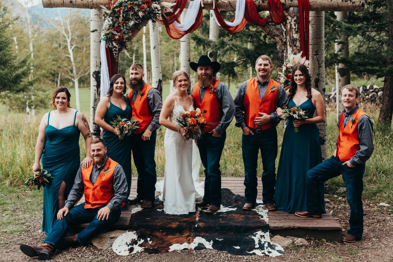 A wedding party poses under a rustic wooden arch with orange and teal color scheme.