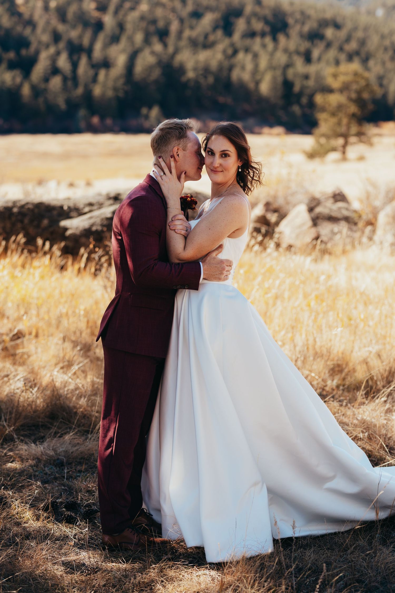 Romantic couple in burgundy suit and white dress embrace in golden mountain meadow.
