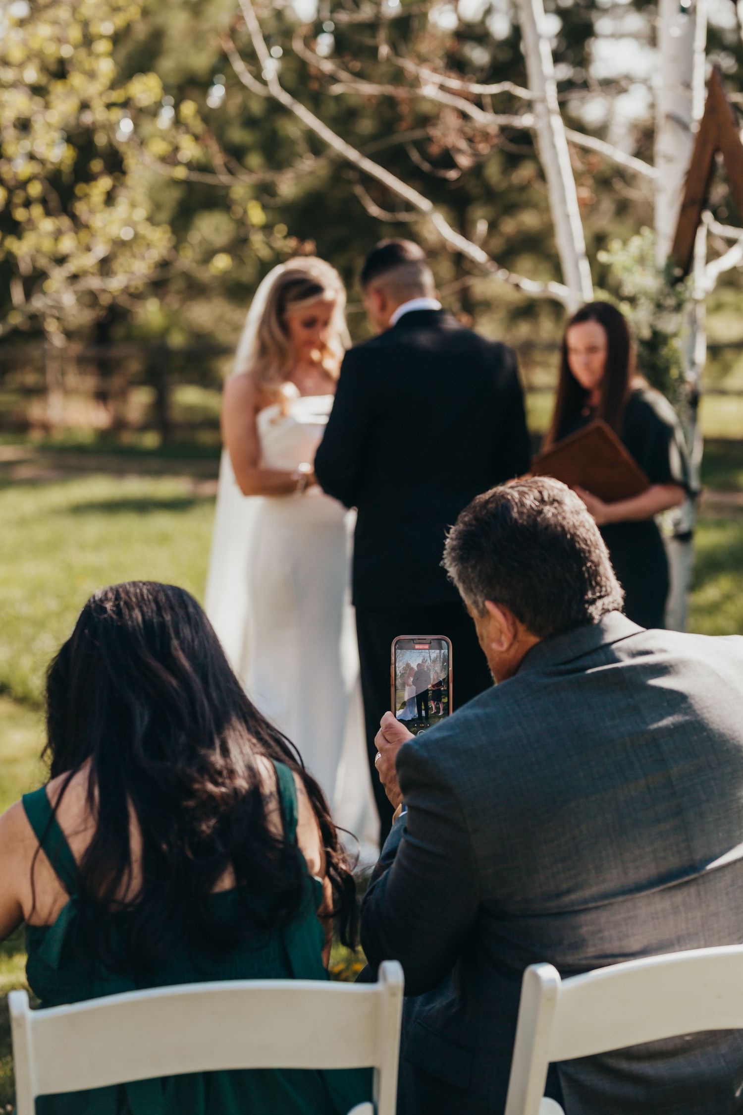 Guests at an outdoor wedding ceremony capturing photos on their phones.